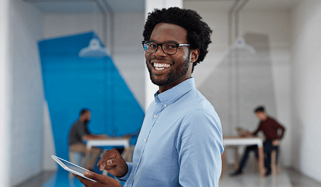 A young Black male, wearing glasses, is carrying a tablet and smiling.